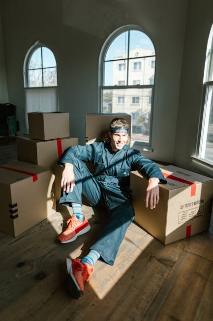 A person sitting on the floor inside a well-lit room with large arched windows, surrounded by several cardboard boxes sealed with red and black packing tape, ready for a home relocation. The individual is dressed in casual clothing, including a blue jumpsuit and bright orange sneakers, and is smiling at the camera with one arm resting on a box. The scene captures a moment during packing and moving preparations, with natural light coming through the windows, highlighting the wooden floor and the boxes stacked behind. This setting reflects the process of furniture transport and packing for a house removal, as handled by Removals Bayswater, ideally suited for a same-day moving service in Bayswater W2.