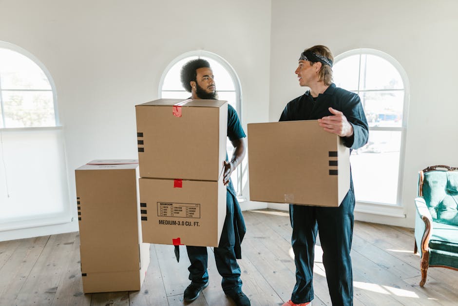 Two movers from Removals Bayswater are inside a well-lit room with large arched windows, engaged in a home relocation process. The mover on the left, with curly hair and a beard, is holding two stacked medium-sized cardboard boxes, labeled with black stripes and red tape, while looking at the other mover. The second mover, with straight hair and wearing a headband, is holding a single cardboard box at chest height and gesturing as if giving instructions or discussing the moving process. The room has a light-colored wooden floor and minimal furnishings, including a vintage-style armchair with a green fabric seat and wooden frame positioned near the window on the right. There are additional cardboard boxes in the background, some stacked on top of bricks, suggesting they are prepared for transport or packing. The natural daylight streaming through the windows creates a bright environment that highlights the efficiency of the packing and loading process, supporting professional house removals and furniture transport services by Removals Bayswater during a home relocation.